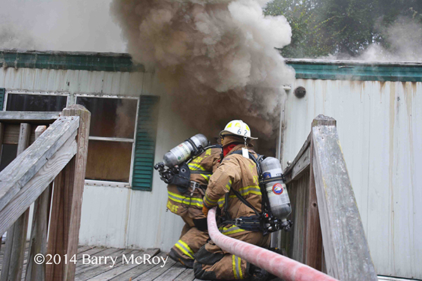 firemen take a hose into a burning mobile home