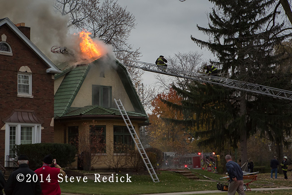 firemen on a ladder with fire through the roof of a house