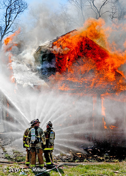 firemen creating a water curtain at a vacant house fully engulfed in flames