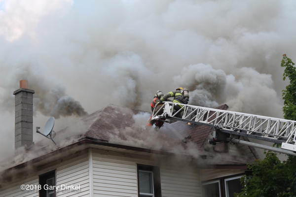 Firefighters at aerial ladder tip on roof with heavy smoke